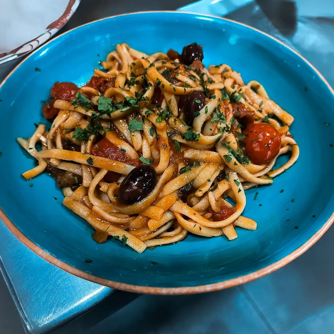 Plato de pasta fresca con tomate, aceitunas negras, verduras salteadas y perejil en un cuenco azul del restaurante Rebeka en Barcelona